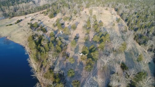 Aerial view of a wooded area with a lake, featuring trees and open fields under a clear sky.