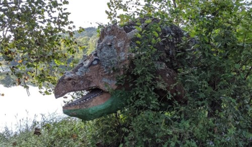 A dinosaur sculpture partially hidden by greenery near a body of water, featuring a textured, colorful face.