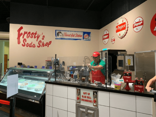 A worker prepares drinks at Frosty's Soda Shop, featuring a soda fountain and a display of ice cream.