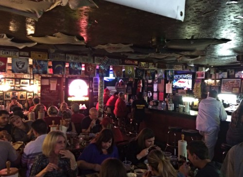 A lively bar scene with patrons dining and watching a game on TV, surrounded by memorabilia and a vibrant atmosphere.