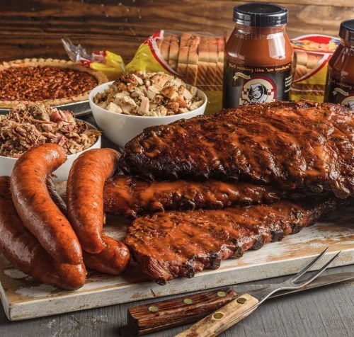 A platter of smoked ribs, sausages, and sides including pie and stuffing, with barbecue sauce jars in the background.