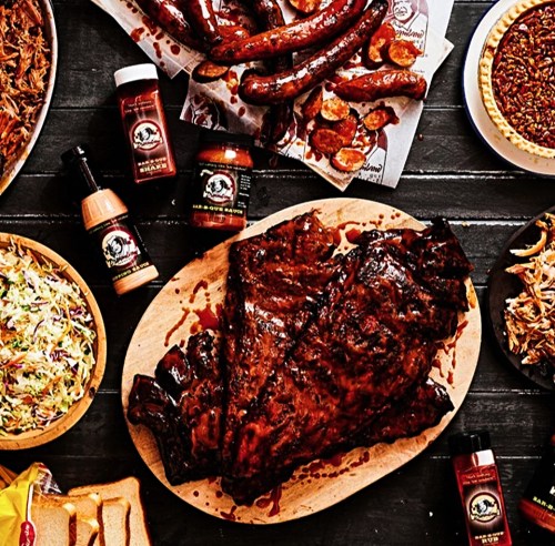A wooden platter with glazed ribs, surrounded by sausages, coleslaw, sauces, and sides on a rustic table.
