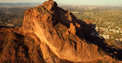 Aerial view of a large rocky mountain with a cityscape in the background, showcasing rugged terrain and greenery.
