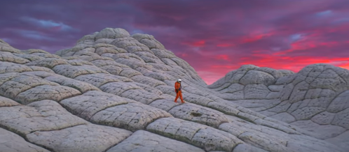 A lone figure in an orange suit walks across textured, rocky terrain under a colorful sunset sky.