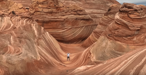 A person stands in a colorful, wavy rock formation, surrounded by towering sandstone cliffs.