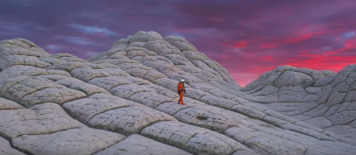 A lone figure in an orange suit stands on textured, rocky terrain under a vibrant, colorful sky at dusk.