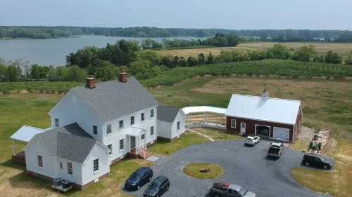 Aerial view of a large white house and red barn near a river, surrounded by green fields and parked vehicles.