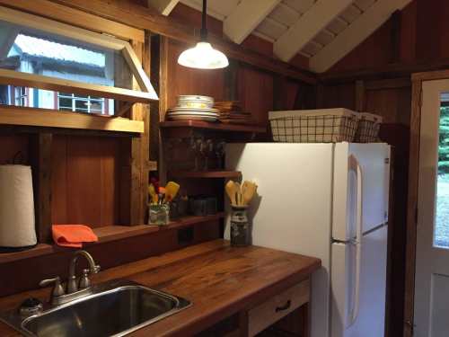 Cozy kitchen with wooden shelves, a white fridge, sink, and rustic decor, illuminated by a hanging light fixture.