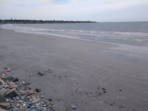 A sandy beach with gentle waves, scattered rocks, and a distant shoreline under a cloudy sky.
