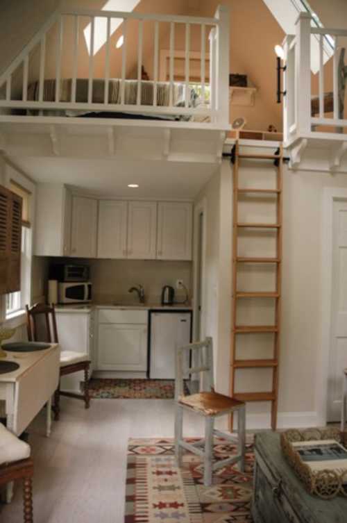 Cozy interior of a small cabin with a loft, kitchenette, and wooden ladder, featuring rustic decor and warm colors.