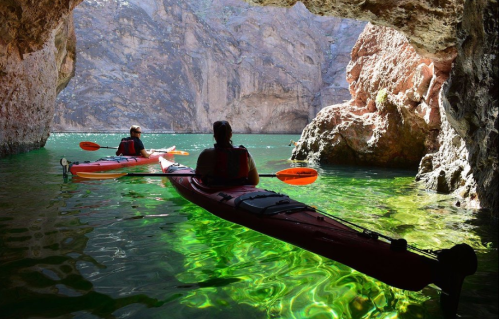 Two kayakers explore a serene cave with vibrant green water and rocky walls reflecting sunlight.