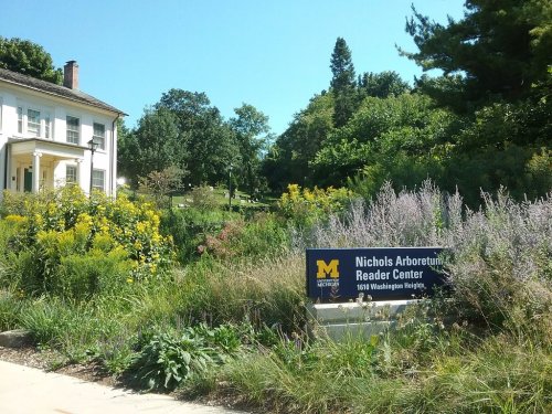 Sign for Nichols Arboretum Reader Center surrounded by lush greenery and colorful flowers on a sunny day.