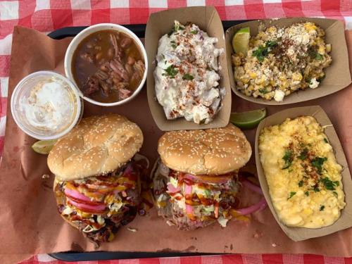 A tray of food featuring two burgers, mac and cheese, corn salad, beans, and a dessert in a cup, all on a checkered tablecloth.