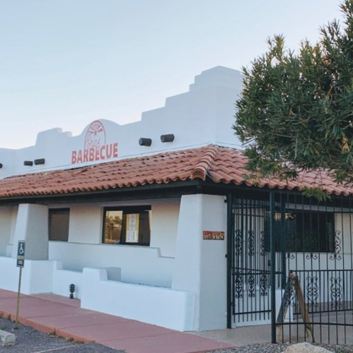 A white building with a red tile roof and a sign that reads "Barbecue," surrounded by desert landscaping.