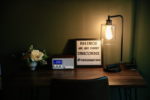A cozy desk with a lamp, a clock radio, a flower arrangement, and a sign that reads "Rhinos are just chubby unicorns."