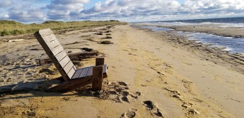 A wooden chair sits on a sandy beach, with gentle waves and clouds in the sky, surrounded by grassy dunes.