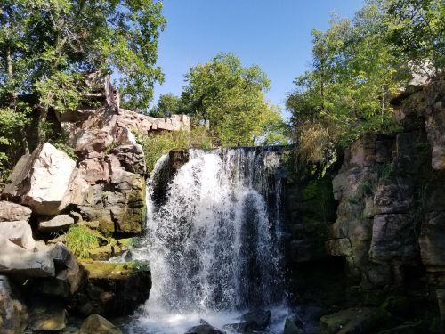 A serene waterfall cascades over rocky terrain, surrounded by lush greenery under a clear blue sky.
