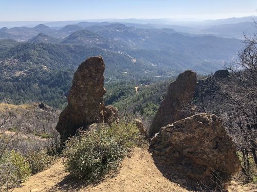 Two large rock formations stand on a hillside, overlooking a vast mountainous landscape with trees and distant hills.