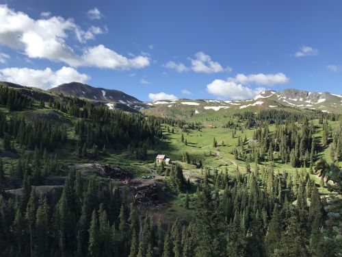 A lush green valley surrounded by mountains, with a small cabin nestled among the trees under a blue sky.