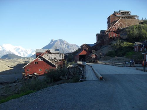 Abandoned red buildings on a hillside with mountains in the background under a clear blue sky.