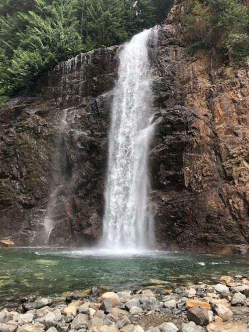A cascading waterfall flows down a rocky cliff into a clear pool, surrounded by lush green trees.