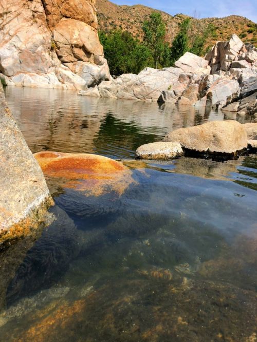 A serene water scene with smooth rocks and clear reflections, surrounded by rugged mountains and greenery.
