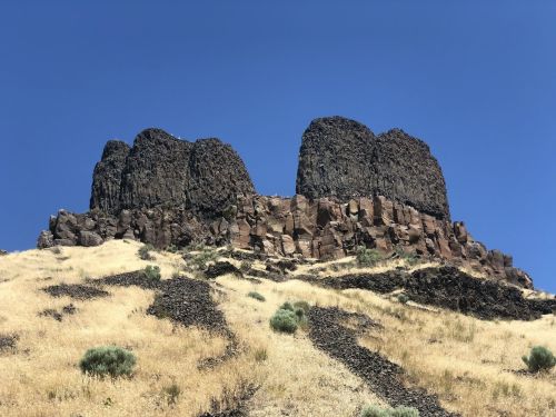 A rocky outcrop with three prominent peaks rises above a dry, grassy hillside under a clear blue sky.