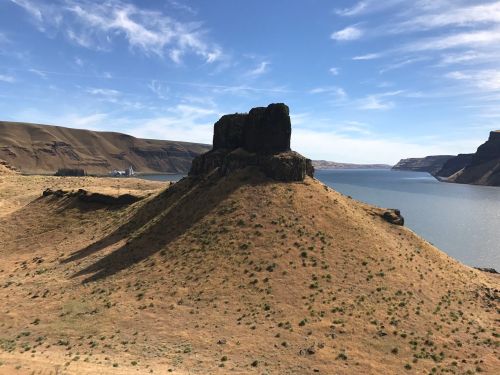 A rocky outcrop rises from a dry landscape, overlooking a calm river with hills in the background under a blue sky.