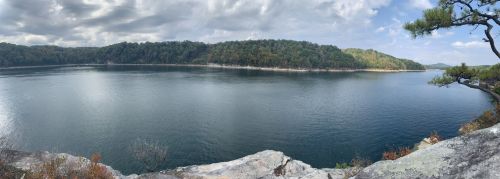 A panoramic view of a calm lake surrounded by lush green hills under a cloudy sky.