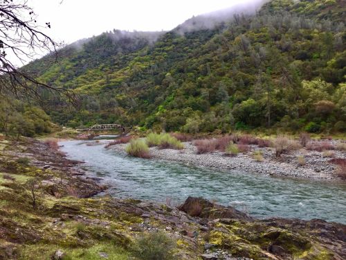 A serene river flows through a lush green landscape, with a bridge in the distance and misty mountains in the background.