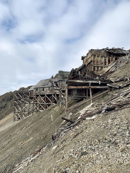 Abandoned wooden structures on a rocky hillside, partially collapsed, under a cloudy sky.