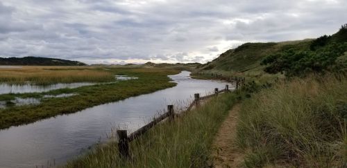 A winding waterway surrounded by grassy banks and dunes under a cloudy sky.