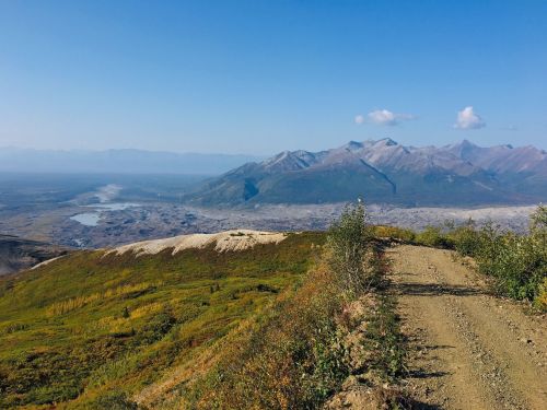 A scenic view of mountains and a valley under a clear blue sky, with a winding dirt path in the foreground.