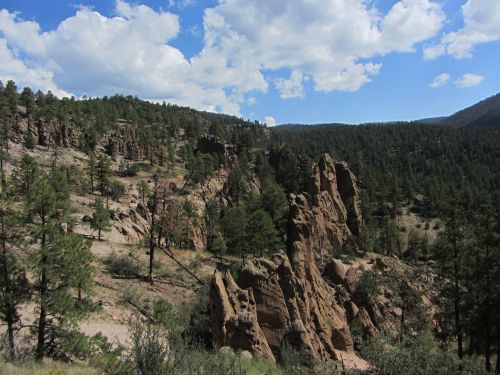 A rocky landscape with towering cliffs, surrounded by pine trees and a blue sky with fluffy clouds.