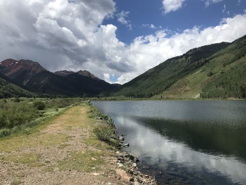 A serene lake surrounded by mountains under a partly cloudy sky, with a gravel path along the water's edge.