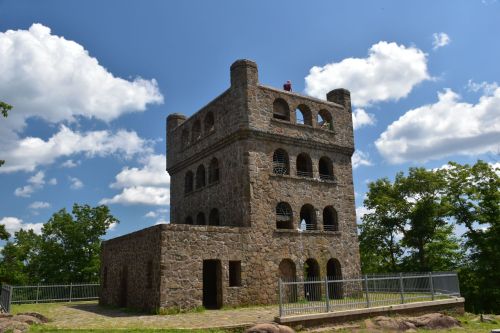 A stone tower with arched windows and a flat roof, surrounded by trees under a blue sky with fluffy clouds.