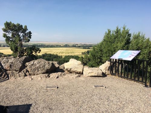 A scenic view from a rocky overlook, featuring fields and distant hills under a clear blue sky.