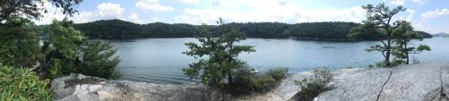 A panoramic view of a calm lake surrounded by lush greenery and rocky shores under a blue sky with scattered clouds.