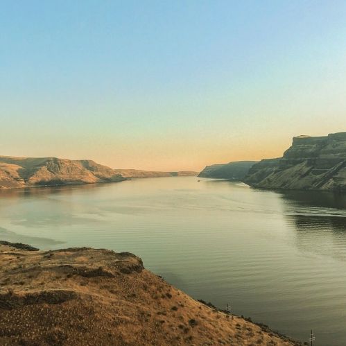 A serene river flows between rocky cliffs under a clear sky, reflecting warm hues of sunset.