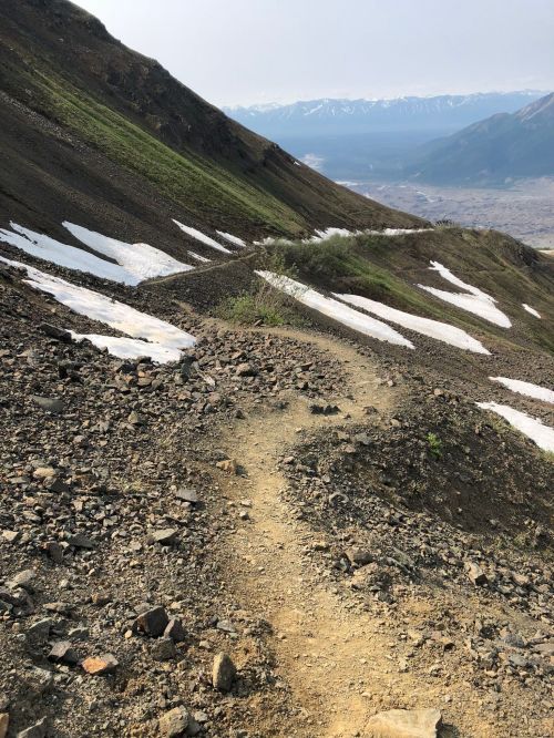 A winding dirt path on a rocky mountainside, with patches of snow and distant snow-capped peaks under a clear sky.