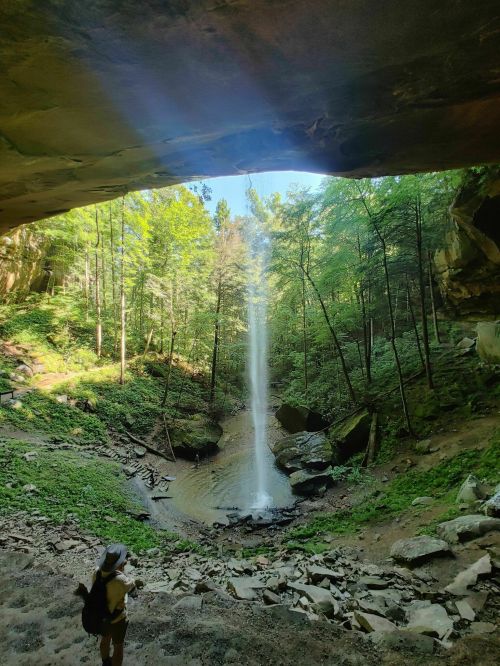 A person stands near a waterfall cascading from a rocky overhang, surrounded by lush green trees and a serene landscape.