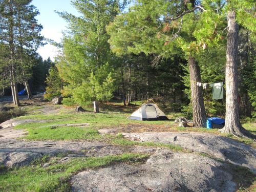 A peaceful campsite with tents set among trees and rocks, featuring hanging clothes and a clear blue sky.
