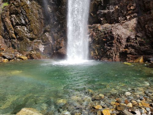 A waterfall cascades into a clear, turquoise pool surrounded by rocky terrain and pebbles.