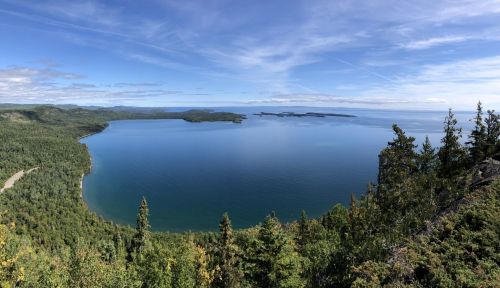 A panoramic view of a serene lake surrounded by lush green forests and distant islands under a clear blue sky.