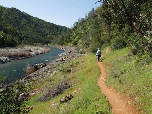 A person walks along a dirt path beside a river, surrounded by lush green hills and trees on a sunny day.