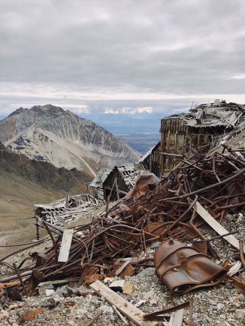 Ruins of a dilapidated building with rusted metal debris, set against a mountainous landscape under a cloudy sky.