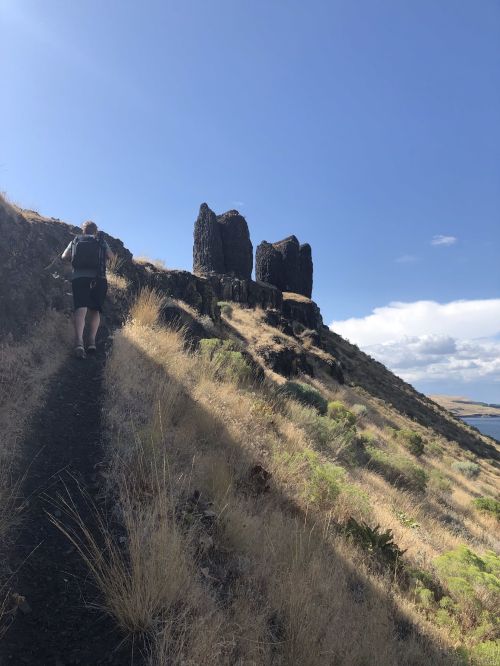 A hiker ascends a rocky trail with two prominent rock formations in the background under a blue sky.