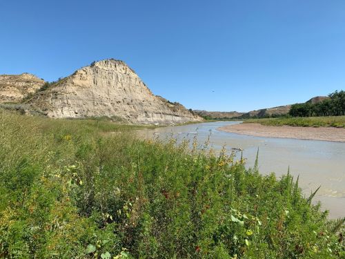 A winding river flows beside a rocky hill, surrounded by lush green vegetation under a clear blue sky.