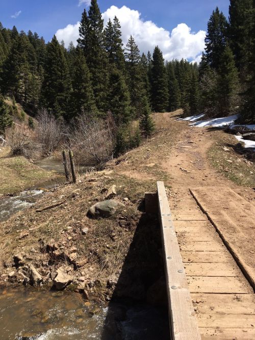 A wooden bridge over a stream on a dirt path surrounded by trees and snow in a mountainous area.
