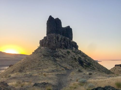 A rocky outcrop rises from a hillside at sunset, with a calm body of water in the background and soft grass in the foreground.
